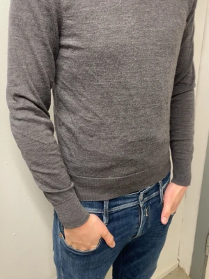 Grå stickad tröja i ullmix - En stilren grå stickad tröja med rund hals och långa ärmar. Tröjan har ribbade muddar vid ärmslut och nederkant, vilket ger en snygg passform. Perfekt att matcha med jeans för en enkel och clean look. Materialet känns mjukt och lätt, troligen en ull- eller bomullsblandning.