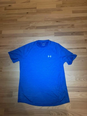 Blå t-shirt från Under Armour - Snygg blå t-shirt från Under Armour med diskret logga på bröstet. T-shirten har korta ärmar och är tillverkad i ett lätt och funktionellt material som passar perfekt för en aktiv livsstil.