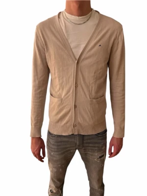 Beige kofta från J.Lindeberg - Snygg beige kofta från J.Lindeberg med diskret logga på bröstet. Koftan har v-ringning, knäppning framtill med tre knappar och två fickor. Tillverkad i mjuk bomull och har ribbade muddar vid ärmslut och nederkant. Perfekt för lager på lager under höst och vår.