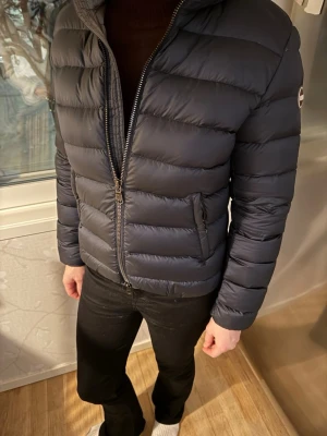 Mörkblå pufferjacka från Colmar - Mörkblå pufferjacka från Colmar med quiltad design, hög krage och två sidofickor med dragkedja. Jackan har en tydlig logotyp på ärmen och är tillverkad i polyester. Perfekt för kalla dagar och ger en clean, modern vibe.