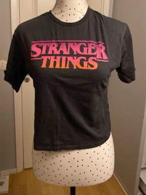 Stranger Things svart croppad t-shirt - Säljer en svart croppad t-shirt med Stranger Things-tryck i rosa och orange på bröstet. T-shirten har rund hals och korta ärmar, perfekt för dig som gillar serien och vill ha en cool och avslappnad stil.