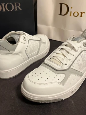 Vita Dior B27 sneakers  - Snygga vita Dior sneakers med grå detaljer och ikoniskt Dior-monogram på sidan. Skorna har rund tå, platt sula och klassisk snörning. Tillverkade i skinn och syntetmaterial för en exklusiv känsla. Perfekta för dig som vill ha något extra till din outfit.