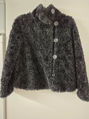 Teddyjacka - Fluffig mörkgrå teddyjacka med hög krage och stora knappar framtill. Jackan har en loose passform och är tillverkad i mjukt syntetmaterial som ger en mysig känsla. Perfekt för dig som vill ha en trendig och bekväm jacka till hösten eller vintern.