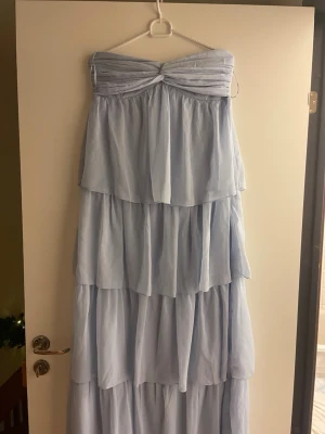 Ljusblå axelbandslös långklänning - Säljer en ljusblå axelbandslös långklänning med flera lager volanger. Klänningen har en twistad detalj vid bysten och är tillverkad i ett tunt, flowy material som ger en romantisk känsla.