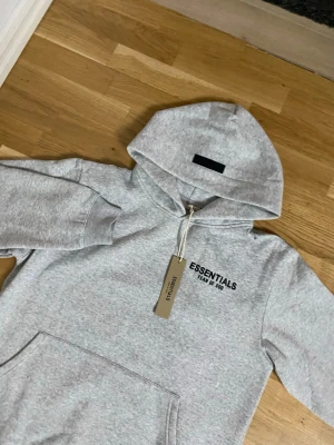 Grå hoodie från Essentials Fear of God - Snygg grå hoodie från Essentials Fear of God med svart logga på bröstet. Klassisk känguruficka framtill och huva med dragsko. Mjuk bomullsblandning som är perfekt för en avslappnad streetwear-look.