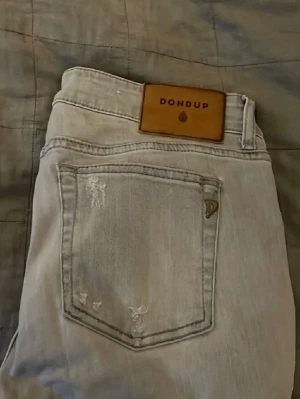 Ljusblå Dondup jeans med slitningar - Säljer ett par ljusblå jeans från Dondup med snygga slitningar på bakfickan och klassisk patch med logga i midjan. Jeansen har en rak passform och är perfekta för en avslappnad stil.