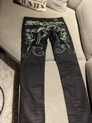 Svarta jeans Kosmo Lupo med drakmotiv - Unika svarta jeans från Kosmo Lupo med stora broderade drakar och grön/vit text på baksidan. Jeansen har raka ben och coola kontrastsömmar. Materialet är jeans med en något glansig finish. Perfekta för dig som vill sticka ut med detaljerad design.