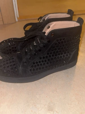 Svarta sneakers med nitar från Louboutin - Säljer ett par svarta high-top sneakers från Christian Louboutin med svarta nitar över hela skon och den klassiska röda sulan. Skorna har snörning och är tillverkade i mocka med detaljer i syntet. Perfekta för dig som vill sticka ut med en edgy stil.