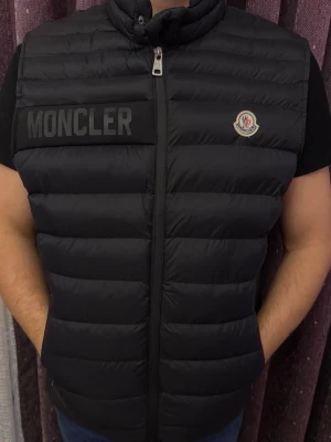 Svart dunväst från Moncler - Svart dunväst från Moncler med hög krage och dragkedja framtill. Västen har Moncler-logga på bröstet och stor Moncler-text på vänster sida. Klassisk quiltad design och lätt vaddering för extra värme.