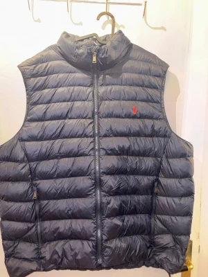 Svart dunväst från Ralph Lauren - Stilren svart dunväst från Ralph Lauren med horisontella sömmar och dragkedja framtill. Västen har två fickor med dragkedja och en liten röd logga på bröstet. Perfekt för lager-på-lager och håller dig varm under kyliga dagar.