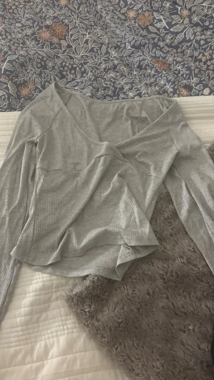 Ljusgrå omlottopp med v-ringning - Säljer en ljusgrå omlottopp med v-ringning och långa ärmar. Toppen är ribbad och har en snygg, enkel design som passar perfekt till jeans eller kjol. Materialet känns mjukt och stretchigt, troligen bomull eller polyesterblandning.