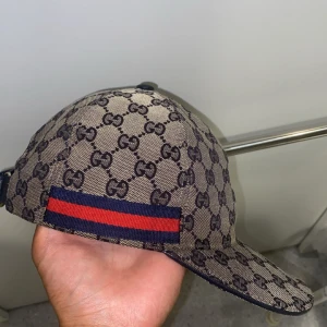 Gucci keps med GG-mönster, M - Snygg keps från Gucci i beige och marinblått med klassiskt GG-monogram över hela kepsen. Har en röd och marinblå rand på sidan och justerbar baksida. Tillverkad i bomullsmix med insida i marinblått tyg. Perfekt för dig som vill sticka ut med lyxig streetstyle. Priset kan diskuteras vid snabb affär 