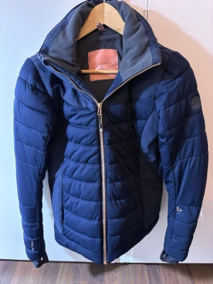 Blå dunjacka  - Snygg blå dunjacka  med quiltad design och dragkedja framtill. Jackan har hög krage, innerficka med dragkedja och diskret logga på ärmen. Perfekt för kalla dagar och har en sportig, modern look.