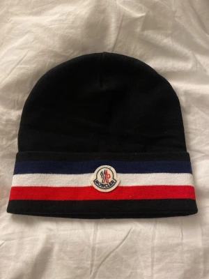 Svart mössa med ränder - Snygg svart mössa med breda blå, vita och röda ränder längst ner samt logga framtill. Klassisk och stilren design i mjukt stickat material som passar perfekt till höst och vinter. OBS mössan sitter lite tajt i början men töjs ut 