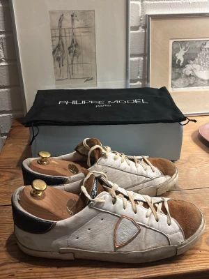 Philippe Model vita sneakers med mockadetaljer - Säljer ett par Philippe Model sneakers i vitt med bruna mockadetaljer på tån och svart hälparti. Skorna har snörning, rund tå och en platt sula med tydlig Philippe Model-logga undertill. Klassisk och stilren design med läder och mocka. Allt i bilder med följer.