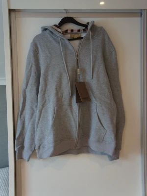 Grå zip hoodie från Burberry - Snygg grå zip hoodie från Barbour med dragkedja framtill, två stora fickor och justerbar huva med snören. Insidan av huvan har ett rutigt tyg som ger en extra detalj. Perfekt för en avslappnad och stilren look. ❗️OBS❗️ - Skriv till mig innan köp! 🙌🏼