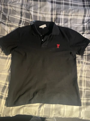 Svart piké från Ami Alexandre Mattiussi - Säljer en svart pikétröja från Ami Alexandre Mattiussi med klassisk krage och två knappar. På bröstet finns Ami:s ikoniska röda hjärtdetalj. Skön och stilren, perfekt till jeans eller chinos. Kan passa om man har M också