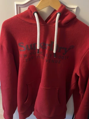 Röd hoodie från Superdry - Snygg röd hoodie från Superdry med tryck på bröstet och vita dragsnören i huvan. Klassisk känguruficka framtill och mjukt, fleeceliknande material på insidan. Perfekt för dig som gillar streetwear och vill ha en bekväm och stilren look.