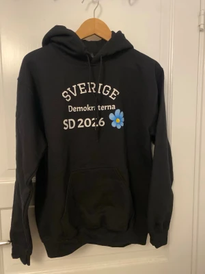 Svart hoodie SD 2026 Sverige - Svart hoodie med huva och magficka. Tryck på bröstet med texten 'Sverige', 'SD 2026', 'Demokraterna' och en blå blomma. Stor blå blomma även på ryggen. Klassisk passform och dragsko i huvan. Perfekt för dig som vill sticka ut.