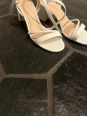 Vita pumps med blockklack från TIAMO GRACE - Stilrena vita pumps från TIAMO GRACE med öppen tå och blockklack. Skorna har tunna remmar över foten och runt vristen med spänne. Perfekta för dig som gillar en enkel men trendig look. Tillverkade i syntetmaterial. använda en halvdag och passar perfekt till studenten, jätte bekväma med 6,5 cm hög klack.