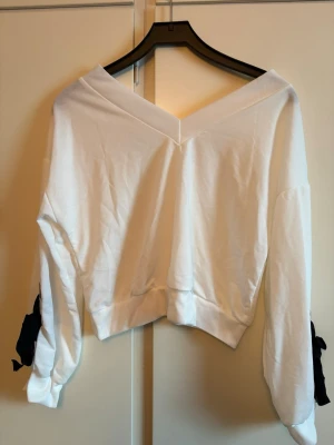 Vit croppad topp med v-ringning - Säljer en vit croppad topp med bred v-ringning både fram och bak. Toppen har långa ärmar med svarta rosetter vid handlederna.