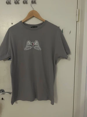 Grå t-shirt från Palm Angels - Snygg grå t-shirt från Palm Angels med tryck av vita vingar och ett hjärta på bröstet. Klassisk rund halsringning och normal passform. Perfekt för dig som gillar streetwear och vill ha något stilrent men ändå unikt.