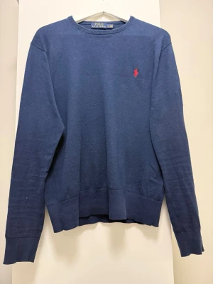 Mörkblå tröja Polo Ralph Lauren Cashmere-blend - Klassisk mörkblå tröja från Polo Ralph Lauren med rund halsringning och röd broderad logga på bröstet. Tröjan har långa ärmar och ribbade muddar vid ärmslut och nederkant. Tröjan är gjord i en cashmere blend