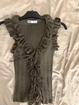 Ribbad volangtop från Zara - Supersnygg ribbad topp från Zara i en mörkgrön ton. Toppen är ärmlös med volangdetaljer längs kanterna och v-ringningen. Perfekt för dig som vill ha en trendig och unik look med extra detaljer.