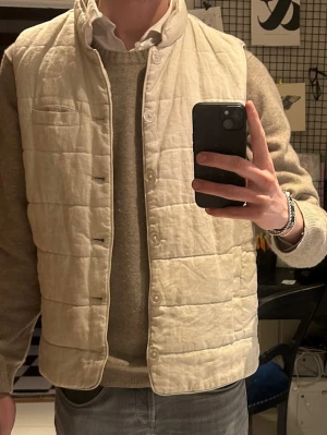 Beige quiltad John Henric linne väst med knappar - Säljer en stilren beige quiltad linneväst från John Henric med ståkrage och knäppning framtill. Västen har två sidofickor och en bröstficka, perfekt för att addera lager till din outfit. Tillverkad i ett mjukt material som känns skönt mot kroppen.