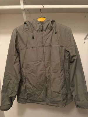 Grå vindjacka med huva och dragkedja - Stilren grå vindjacka med hel dragkedja framtill och justerbar huva med snörning. Jackan har en enkel design och är perfekt för blåsiga dagar. Tillverkad i syntetmaterial och har långa ärmar för extra skydd.