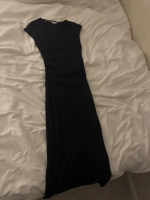 Svart långklänning med öppen rygg - En svart långklänning med tight passform och öppen rygg. Klänningen har korta ärmar och en stilren, minimalistisk design. Perfekt för dig som gillar en enkel men elegant look. Prislappen är kvar men själva lappen är borta, se bild 3.