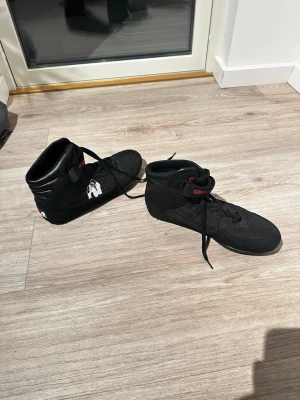 Svarta Gorilla Wear träningsskor - Säljer ett par svarta träningsskor från Gorilla Wear med högt skaft och kardborreband. Skorna har vadderad häl, snörning och detaljer i rött och vitt, inklusive Gorilla Wear-loggan på sidan och hälen. Perfekta för gym och styrketräning.