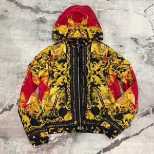 Versace Jeans Couture vindjacka  - Säljer en Versace Jeans Couture vindjacka med ikoniskt barockmönster i guld och svart, samt röda detaljer. Jackan har huva, två fickor med dragkedja och snygga snörningar. Materialet är lätt polyester och jackan har en stor rund dragkedja framtill med Versace-logga. Storlek IT:40 vilket motsvarar S. Retail: 4800 kr. Äkthetskontroll kan man göra på själva hemsidan och använda koden som finns i jackans lappar (CLG) OBS! PRIS GÅR ATT DISKUTERA.
