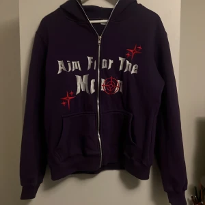 Lila hoodie Aim For The Moon M - Snygg lila hoodie från Aim For The Moon i storlek M. Tröjan har huva, dragkedja och fickor framtill. Tryck i vitt och rött med texten 'Aim For The Moon' och coola grafiska detaljer. Perfekt för dig som gillar streetwear och vill sticka ut.