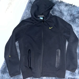 Svart Nike NOCTA vindjacka med huva - Svart vindjacka från Nike NOCTA med huva och hel dragkedja. Jackan har gula detaljer, inklusive NOCTA-logga och Nike Swoosh på bröstet. Två fickor med dragkedja framtill och stilren design. Perfekt för streetwear och chill dagar.
