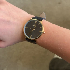 Svart och guld klocka från MEMA - Stilren klocka från MEMA med svart urtavla och guldiga detaljer. Rund boett i guldtonad metall och romerska siffror. Armbandet är blått med krokodilpräglat mönster och spänne i silver. Baksidan är i rostfritt stål och klockan är vattentålig.