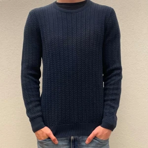 Mörkblå stickad tröja från J.Lindeberg - Säljer en mörkblå stickad tröja från J.lindeberg med snyggt strukturmönster. Tröjan har rund halsringning, ribbade muddar och lång ärm. Perfekt för dig som gillar stilrena och klassiska plagg med lite extra detalj. Har blivit solskadad men synns knappt när man har den på sig