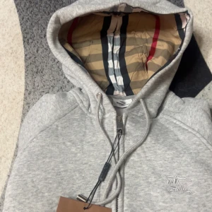 Grå Burberry hoodie med dragkedja - Säljer en grå hoodie från Burberry med klassiskt rutigt foder i huvan och broderad logga på bröstet. Tröjan har snörning i huvan, dragkedja framtill och ribbade muddar. Perfekt för dig som gillar stilrena och lyxiga detaljer.