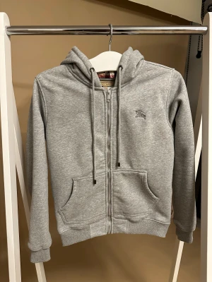 Grå Burberry hoodie med rutig huva - Snygg grå hoodie från Burberry med rutig insida på huvan. Tröjan har använts ett fåtal gånger. Säljer den pågrund av att den aldrig används. Vid frågor hör av er.