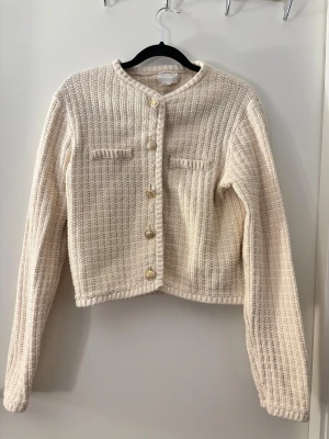 Beige stickad cardigan jacka med guldknappar - Säljer en beige stickad cardigan jacka med snygga guldknappar framtill och två dekorativa fickor på bröstet. Jackan har en croppad passform och är långärmad med rund hals. Perfekt för dig som gillar en clean och stilren look.