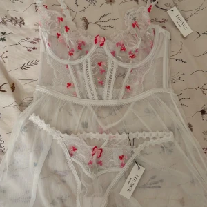 Vit blommig korsett från Lounge - Superfin vit korsett från Lounge med transparent mesh och broderade rosa och röda blommor. Detaljerad spets längs kanterna och en söt rosett framtill. Axelbanden är justerbara och hela toppen har en romantisk vibe med blommiga broderier och spets. Trosorna är sålda så endast nattlinnet kvar!!