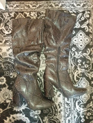 Bruna boots med ormskinnsmönster - Snygga bruna boots från Seven Dials med ormskinnsmönster och hög klack. Skorna har spetsig tå, dragkedja på sidan och är tillverkade i syntetmaterial. Perfekta för dig som vill sticka ut med ett coolt statement. Tror att storleken är 38, jag har storlek 39 och de blir riktigt tajta när jag provade de. De är endast provade ej använda.