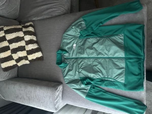 Grön vindjacka från The North Face - *Oanvänd* (pris kan diskuteras) Snygg grön vindjacka från The North Face med två olika gröna nyanser och hel dragkedja framtill. Jackan har lång ärm, hög krage och logga på både bröstet och ryggen. Materialet är lätt och syntetiskt, perfekt för utomhusaktiviteter och chill dagar. Köpt i jd sport nypris 1000kr 