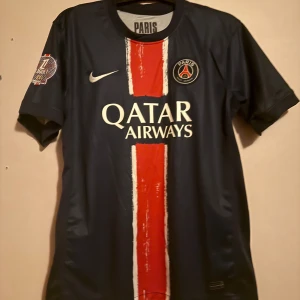 PSG Dembele 10 Nike fotbollströja - Säljer en officiell Paris Saint-Germain fotbollströja från Nike med O. Dembélé och nummer 10 på ryggen. Tröjan är mörkblå med en bred röd och vit rand i mitten, PSG-märke på bröstet och Qatar Airways som sponsor. Korta ärmar och klassisk passform.