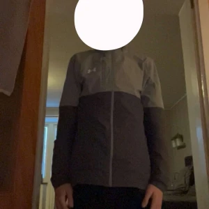 Grå vindjacka från Under Armour - Säljer en snygg tvåtonad grå vindjacka från Under Armour i storlek M. Jackan har huva, hel dragkedja framtill och långärmade ärmar. Perfekt för träning eller när du vill ha en sportig look. Materialet är lätt och känns skönt mot kroppen.