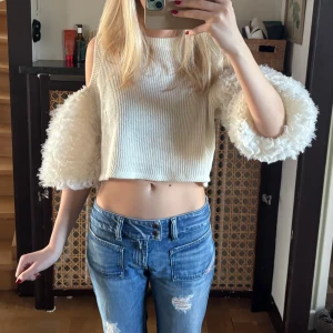 Vit croppad topp med fluffiga ärmar - Säljer en vit croppad topp med ribbad struktur och unika fluffiga långärmar. Toppen har öppen axel-design och är tillverkad i stickat material med pälsdetaljer på ärmarna. Perfekt för dig som vill sticka ut med en cool och trendig look. Se defekt - sista bilden!