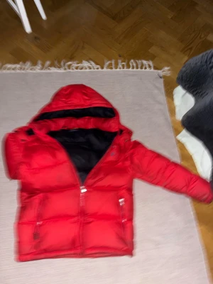 Röd dunjacka från Polo Ralph Lauren - Riktigt fet röd dunjacka från Polo Ralph Lauren med svart innerfoder och huva. Jackan har dragkedja framtill, två sidofickor med dragkedja och en bröstficka. Perfekt för kalla dagar och har en klassisk pufferstil som sticker ut. Storlek XL barn
