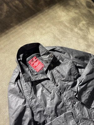 Svart vindjacka från J&J Vintage - Svart vindjacka från Jack & Jones Vintage med hög krage, dragkedjor och knappar framtill. Jackan har flera fickor, axelklaffar och detaljerade sömmar. Materialet är syntet och jackan har en cool, urban vibe som passar perfekt till streetwear.