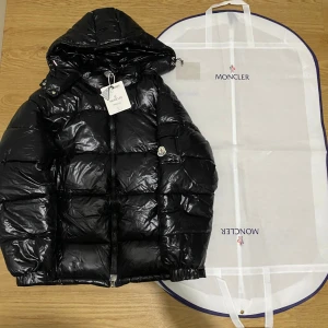 Svart pufferjacka från Moncler  - Svart pufferjacka från Moncler i storlek L med glansig finish och klassisk Moncler-logga på ärmen. Jackan har huva, dragkedja framtill och coolt serietidningsfoder på insidan. Perfekt för dig som vill ha en varm och snygg jacka med streetvibe.
