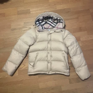 Beige pufferjacka från Burberry - Säljer en beige pufferjacka från Burberry med avtagbara ärmar så den kan användas som väst. Jackan har en huva med klassiskt rutigt Burberry-foder i svart, vit och rosa. Två fickor med dragkedja och knappar framtill. Perfekt för kalla dagar.
